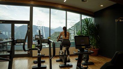 Frau trainiert auf Heimtrainer in Fitnessraum mit Bergblick durch große Fenster