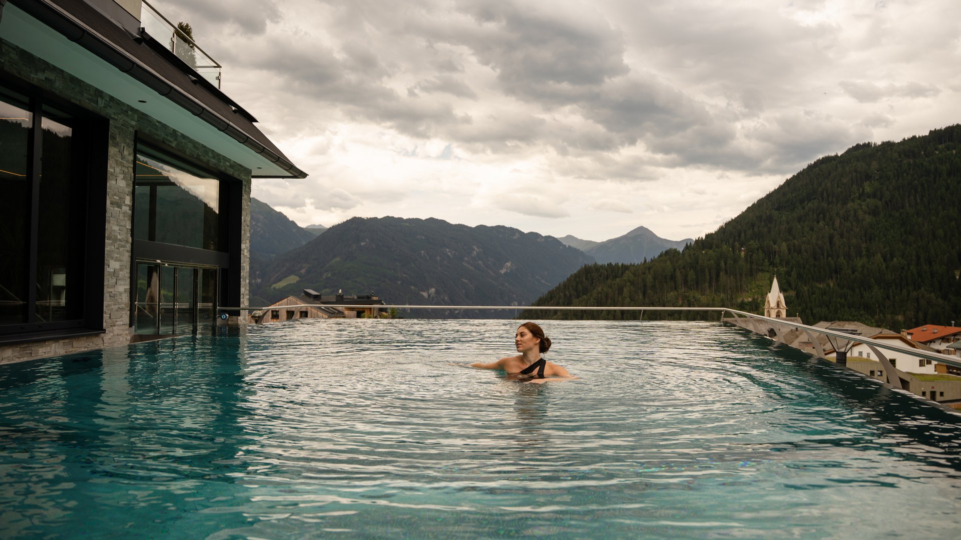 Frau schwimmt im Infinity-Pool mit Bergblick und bewölktem Himmel